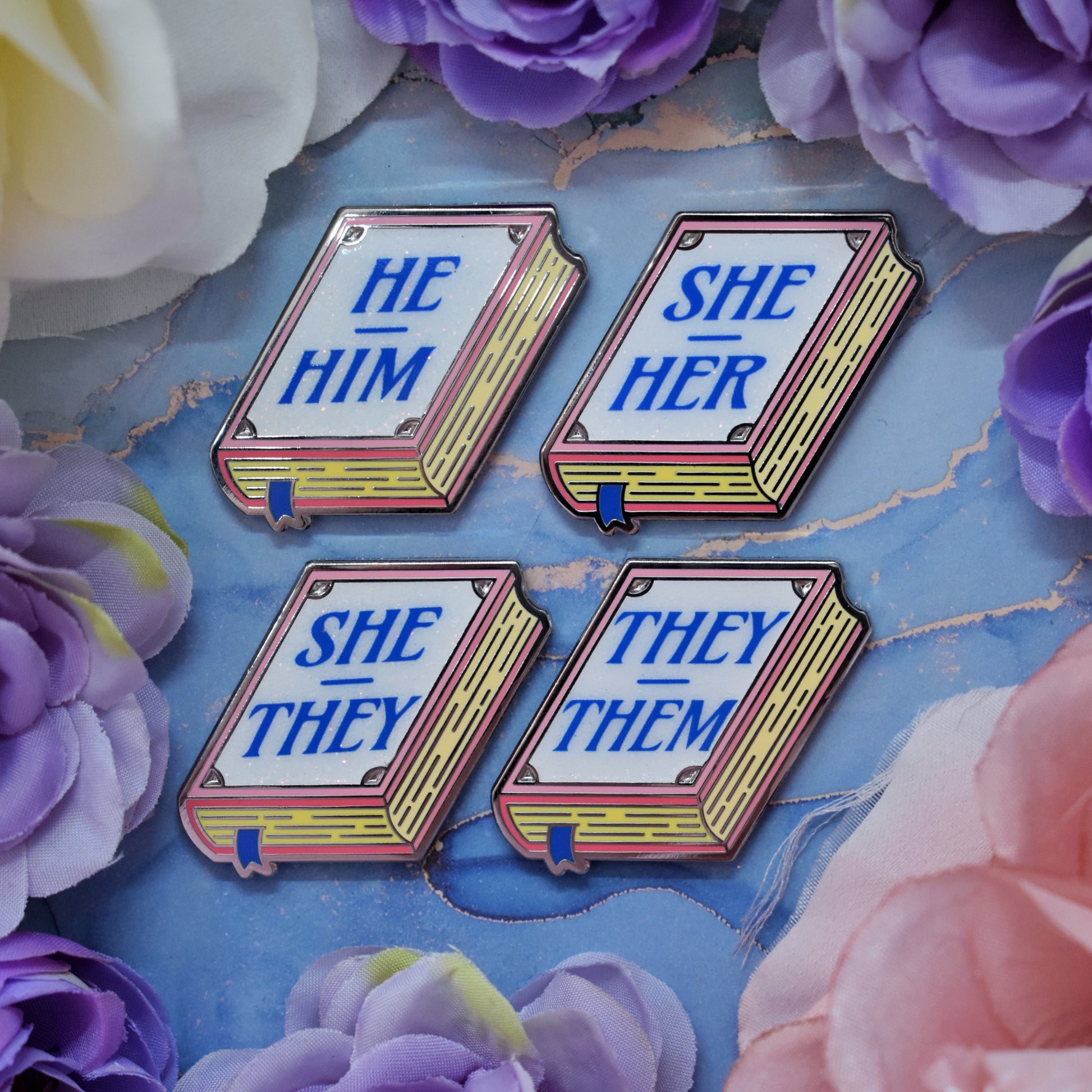 Four pink, white, yellow book shaped enamel pins with text 'HE HIM', 'SHE HER', 'SHE THEY', 'THEY THEM' in blue on a watercolour background with a floral border.