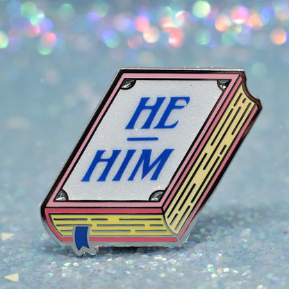 Book shaped enamel pin in silver metal, featuring pink, yellow and white glitter accents with 'HE - HIM' in a midtone blue. Background is a glittery icey blue.