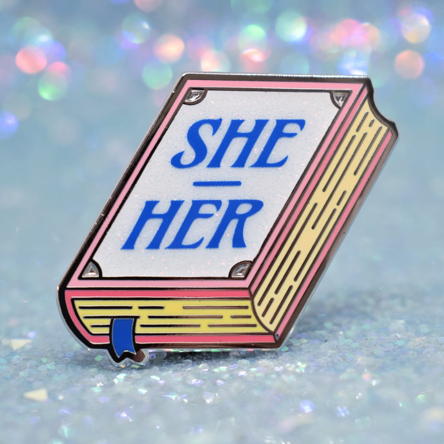 Book shaped enamel pin in silver metal, featuring pink, yellow and white glitter accents with 'SHE - HER' in a midtone blue. Background is a glittery icey blue.