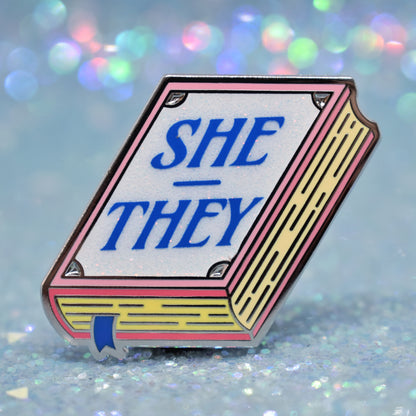 Book shaped enamel pin in silver metal, featuring pink, yellow and white glitter accents with 'SHE - THEY' in a midtone blue. Background is a glittery icey blue.