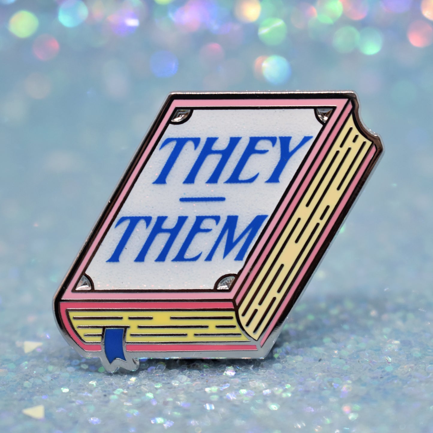 Book shaped enamel pin in silver metal, featuring pink, yellow and white glitter accents with 'THEY - THEM' in a midtone blue. Background is a glittery icey blue.