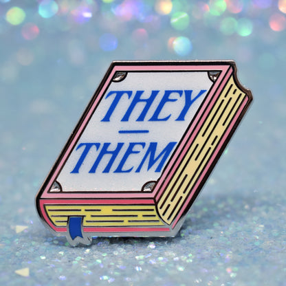 Book shaped enamel pin in silver metal, featuring pink, yellow and white glitter accents with 'THEY - THEM' in a midtone blue. Background is a glittery icey blue.