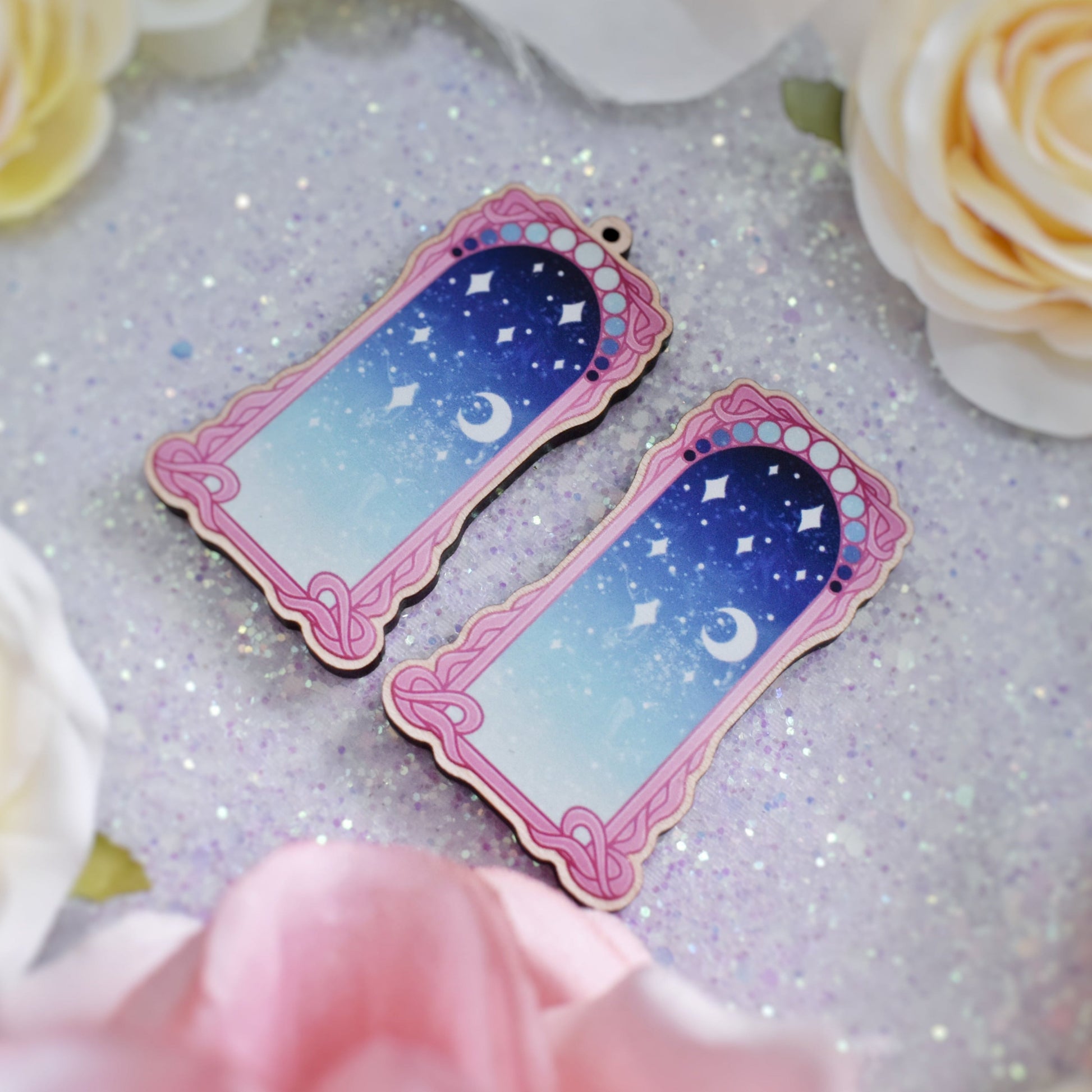 2 wooden charms, 1 with a loop at the top, 1 without next to each other at an angle on a sparkly background. Designs have pink detailed frame. Gradient colour pale teal through to blue with stars and a moon inside frame.