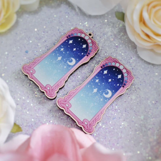 2 wooden charms, 1 with a loop at the top, 1 without next to each other at an angle on a sparkly background. Designs have pink detailed frame. Gradient colour pale teal through to blue with stars and a moon inside frame.