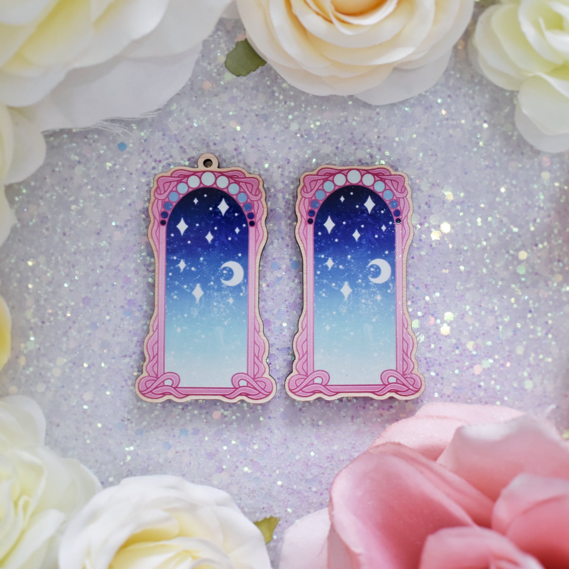 2 wooden charms, 1 with a loop at the top, 1 without next to each other on a sparkly background. Designs have pink detailed frame. Gradient colour pale teal through to blue with stars and a moon inside frame.