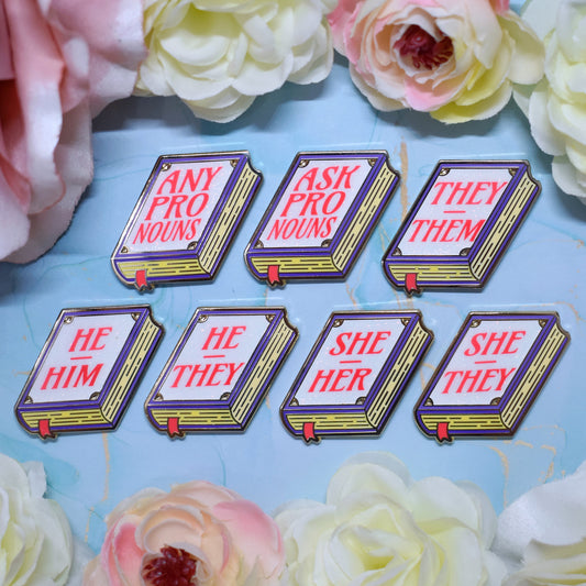 Seven purple, white, yellow book shaped enamel pins with text 'ANY PRONOUNS' 'ASK PRONOUNS' 'HE HIM', 'HE THEY' 'SHE HER', 'SHE THEY', 'THEY THEM' in pink on a watercolour background with a floral border.