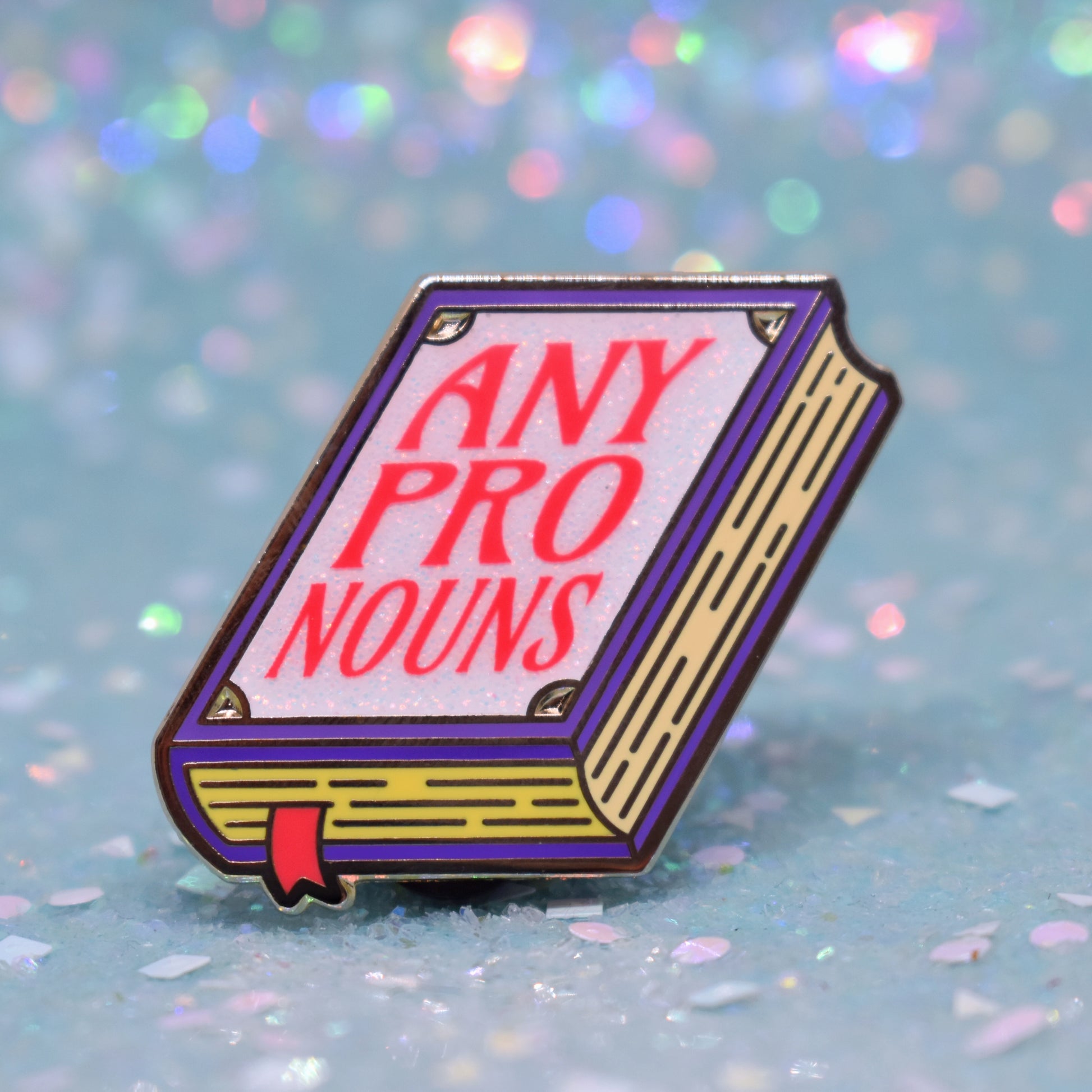 Book shaped enamel pin in gold metal, featuring purple, yellow and white glitter accents with 'ANY PRONOUNS' in a bright red/pink. Background is a glittery icy blue.