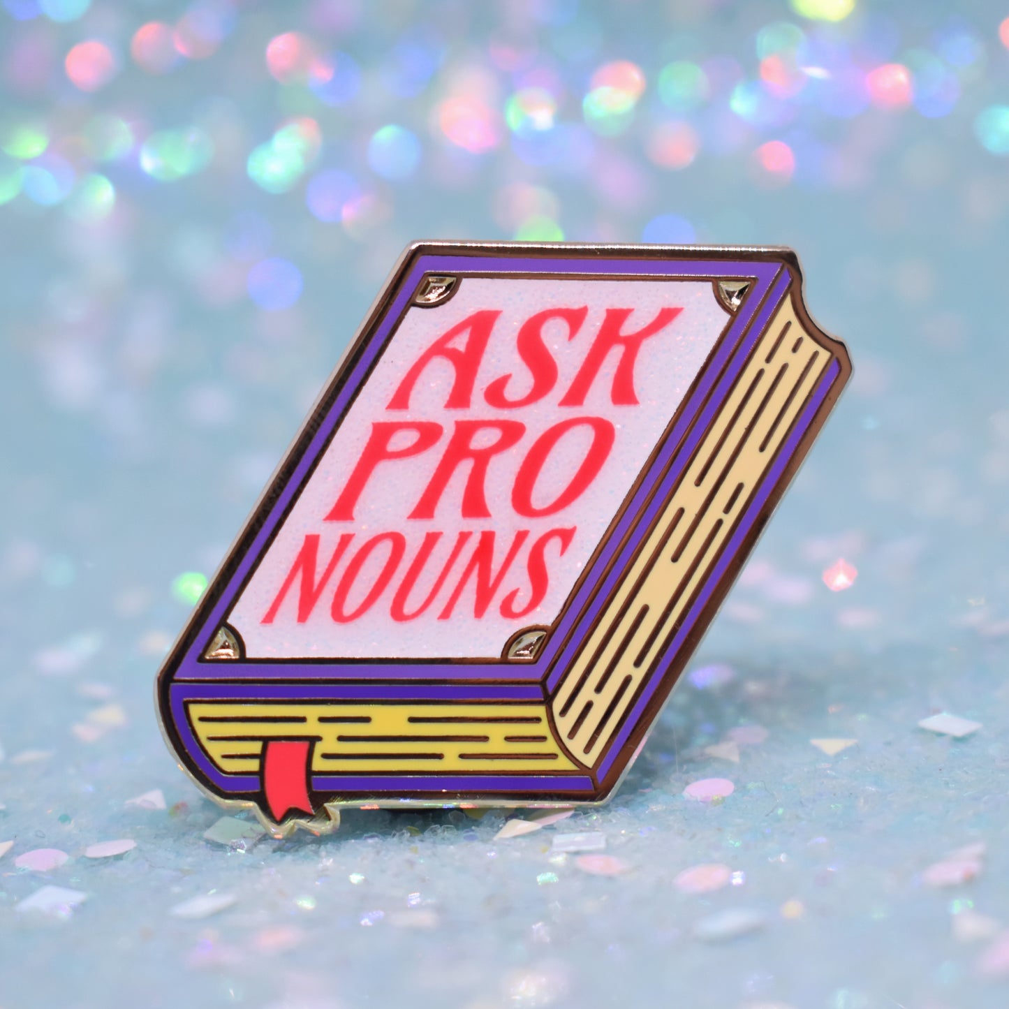 Book shaped enamel pin in gold metal, featuring purple, yellow and white glitter accents with 'ASK PRONOUNS' in a bright red/pink. Background is a glittery icy blue.