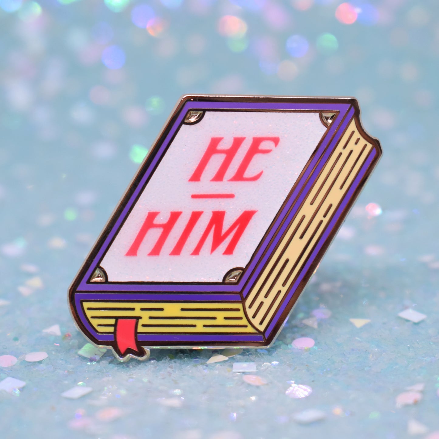 Book shaped enamel pin in gold metal, featuring purple, yellow and white glitter accents with 'HE - HIM' in a bright red/pink. Background is a glittery icy blue.