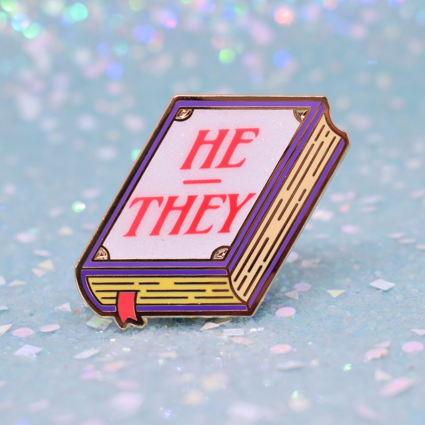 Book shaped enamel pin in gold metal, featuring purple, yellow and white glitter accents with 'HE - THEY' in a bright red/pink. Background is a glittery icy blue.