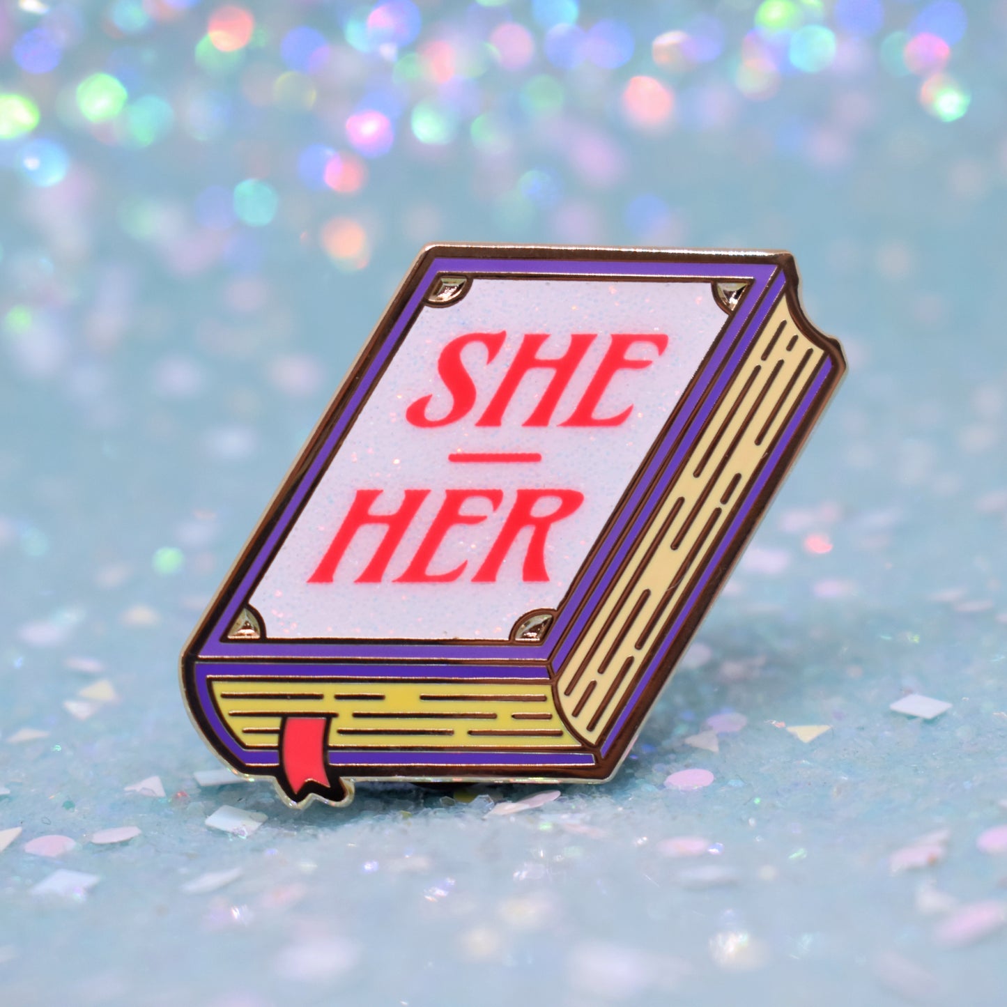 Book shaped enamel pin in gold metal, featuring purple, yellow and white glitter accents with 'SHE - HER' in a bright red/pink. Background is a glittery icy blue.