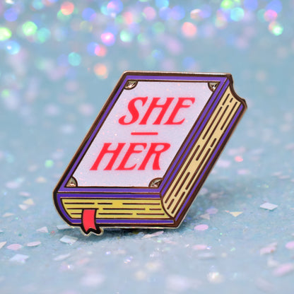 Book shaped enamel pin in gold metal, featuring purple, yellow and white glitter accents with 'SHE - HER' in a bright red/pink. Background is a glittery icy blue.
