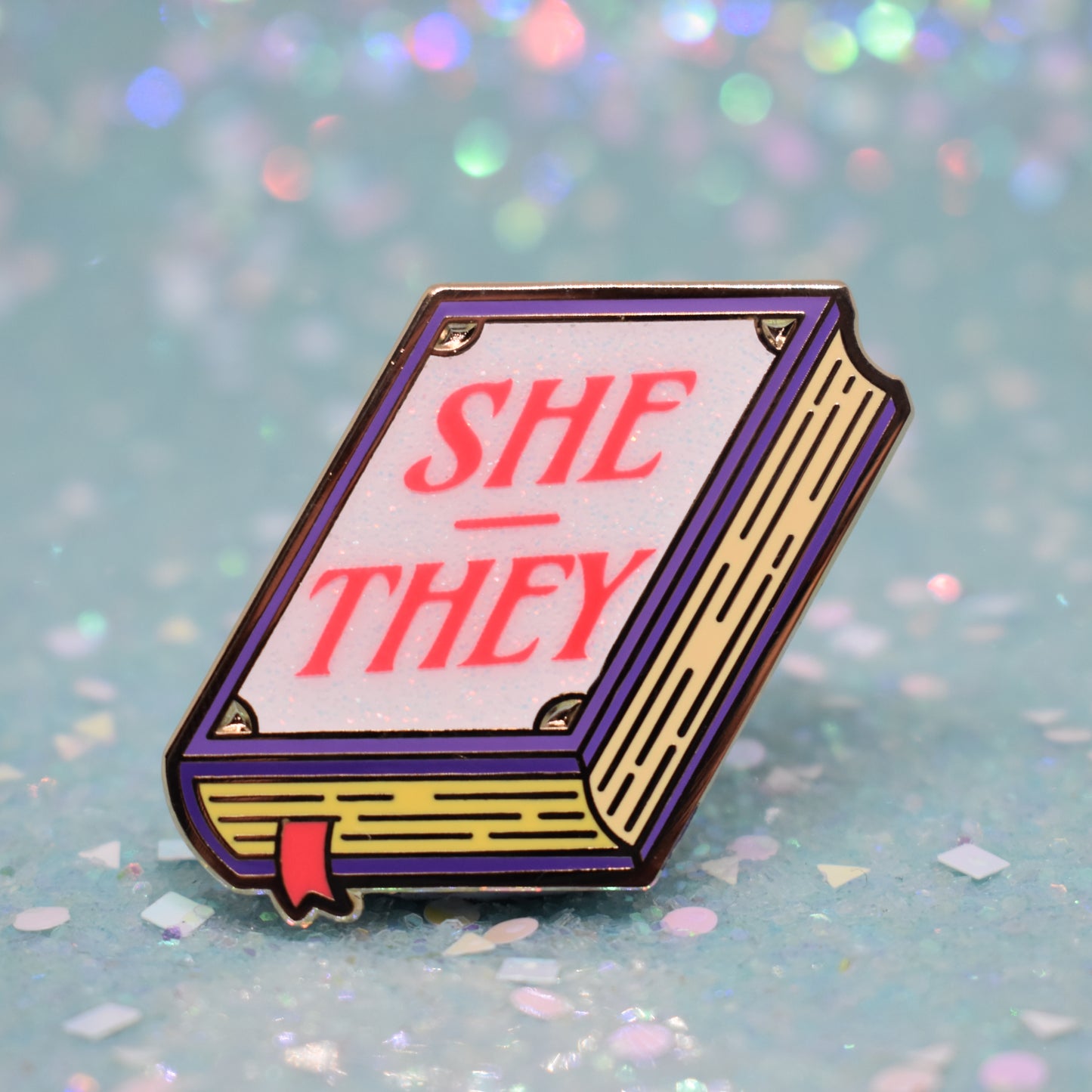 Book shaped enamel pin in gold metal, featuring purple, yellow and white glitter accents with 'SHE - THEY' in a bright red/pink. Background is a glittery icy blue.