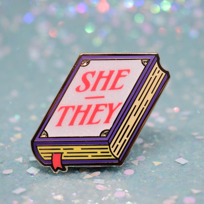 Book shaped enamel pin in gold metal, featuring purple, yellow and white glitter accents with 'SHE - THEY' in a bright red/pink. Background is a glittery icy blue.