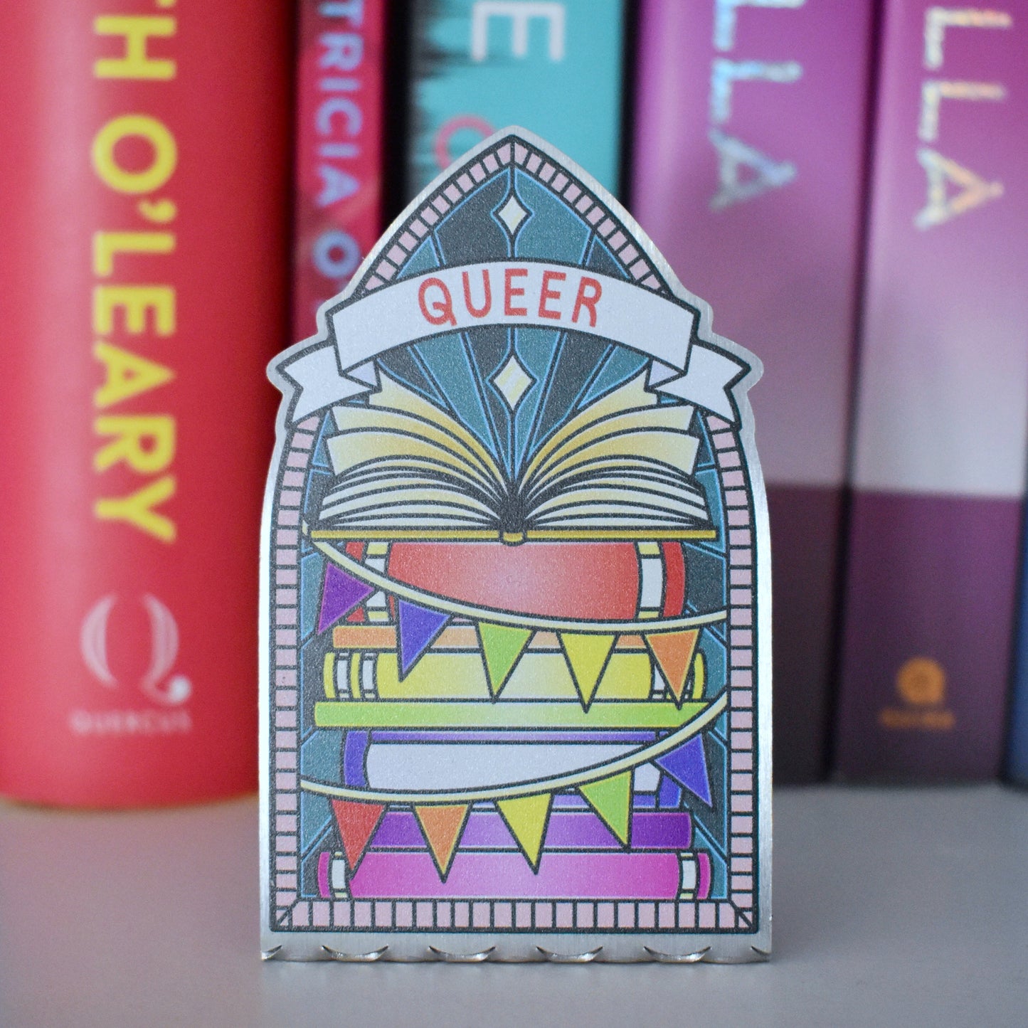 Decorative stainless steel standee with 'Queer' design on in the style of a stained glass window with a row of books behind.