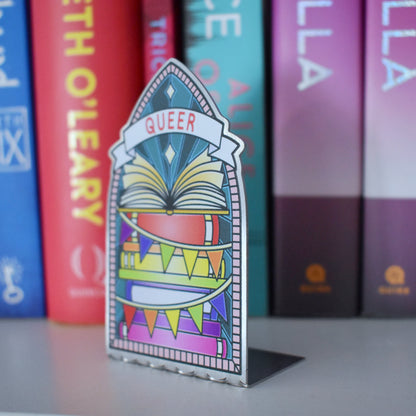 Decorative stainless steel standee with 'Queer' design on in the style of a stained glass window at an angle to see it is an L shape with a row of books behind.