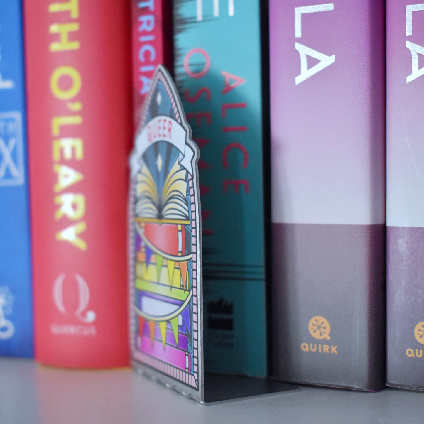 Decorative stainless steel standee with 'Queer' design on in the style of a stained glass window at an angle partically pushed under the row of books, to see it is an L shape that fits under books.