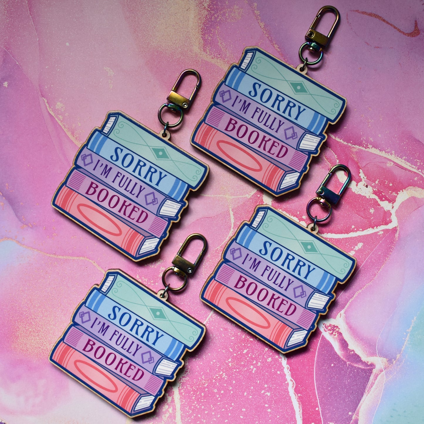 Four wooden keyrings in a slanted diamond shape, with a colourful book stack design with detailed spines and 'Sorry I'm Fully Booked' text. On a colourful watercolour style background.