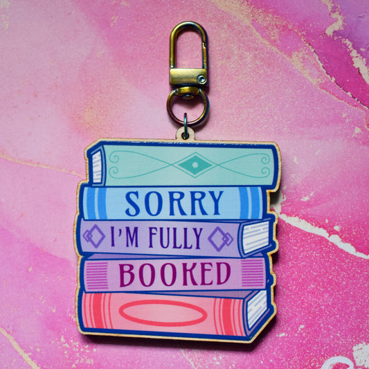 Wooden keyring with colourful book stack design with detailed spines and 'Sorry I'm Fully Booked' text. On a colourful watercolour style background.