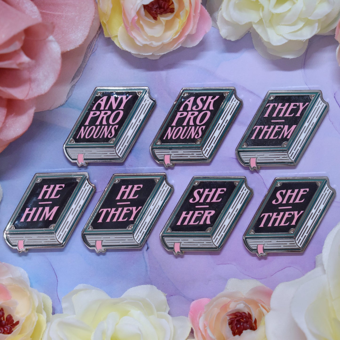 Seven teal, white, black book shaped enamel pins with text 'ANY PRONOUNS' 'ASK PRONOUNS' 'HE HIM', 'HE THEY' 'SHE HER', 'SHE THEY', 'THEY THEM' in pink on a watercolour background with a floral border.