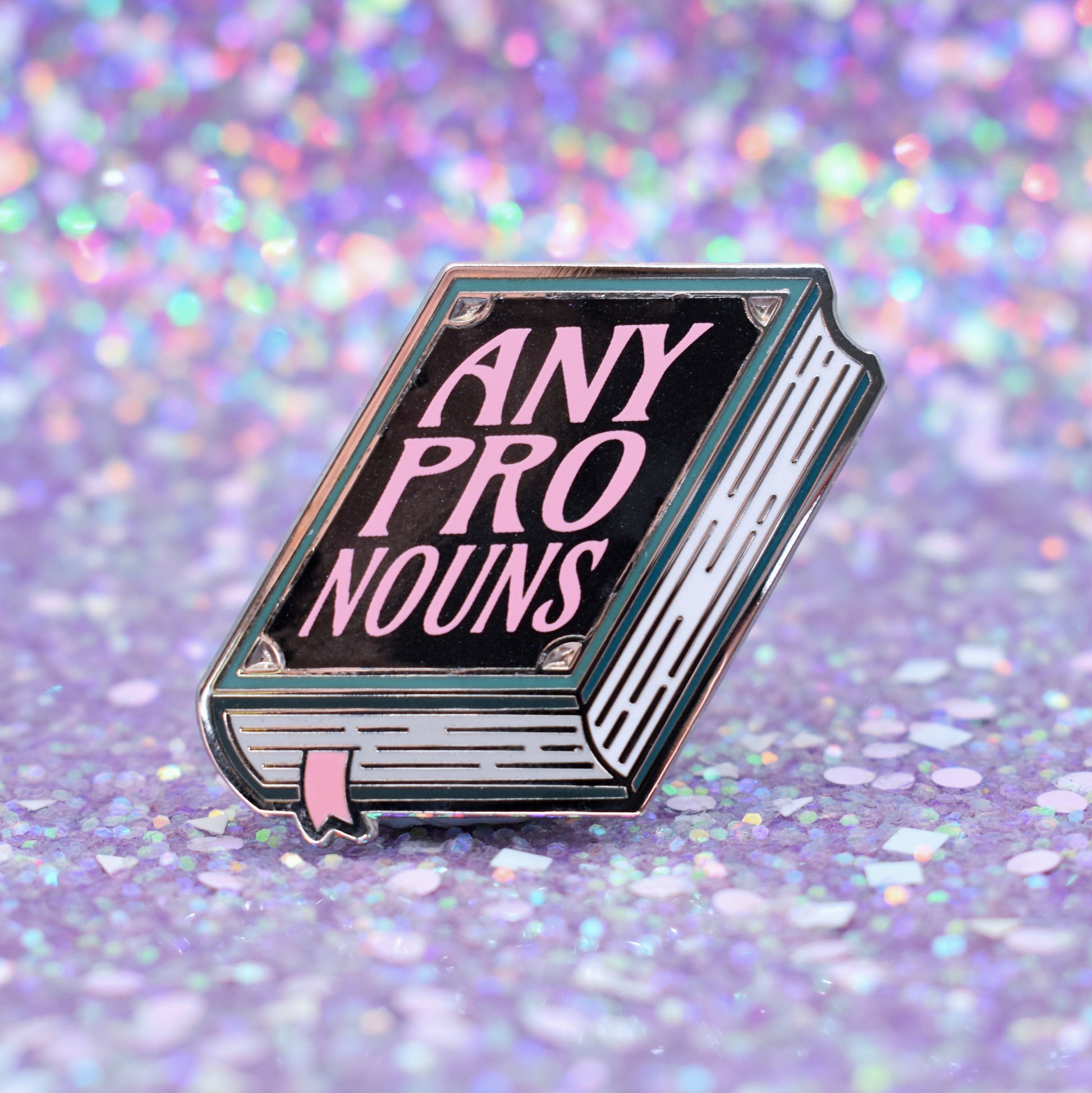 Book shaped enamel pin in silver metal, featuring teal, white and black shimmer accents with 'ANY PRONOUNS' in a light pink. Background is a glittery lilac.