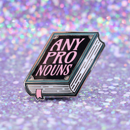 Book shaped enamel pin in silver metal, featuring teal, white and black shimmer accents with 'ANY PRONOUNS' in a light pink. Background is a glittery lilac.