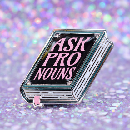Book shaped enamel pin in silver metal, featuring teal, white and black shimmer accents with 'ASK PRONOUNS' in a light pink. Background is a glittery lilac.