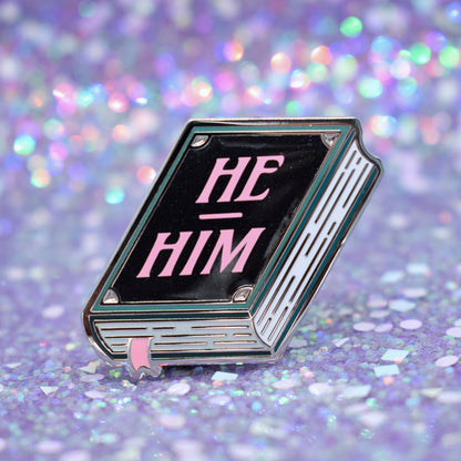 Book shaped enamel pin in silver metal, featuring teal, white and black shimmer accents with 'HE - HIM' in a light pink. Background is a glittery lilac.