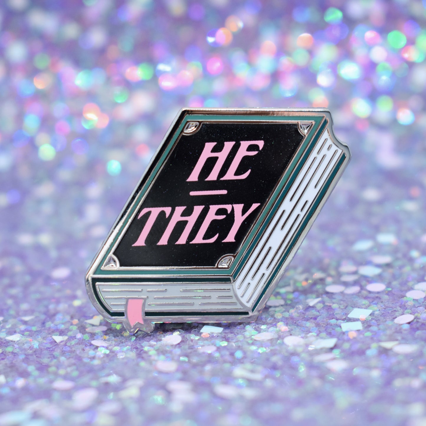 Book shaped enamel pin in silver metal, featuring teal, white and black shimmer accents with 'HE - THEY' in a light pink. Background is a glittery lilac.