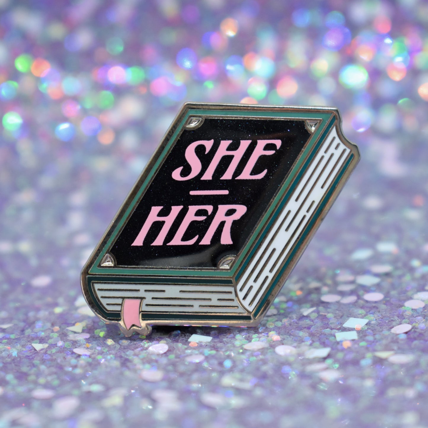 Book shaped enamel pin in silver metal, featuring teal, white and black shimmer accents with 'SHE - HER' in a light pink. Background is a glittery lilac.