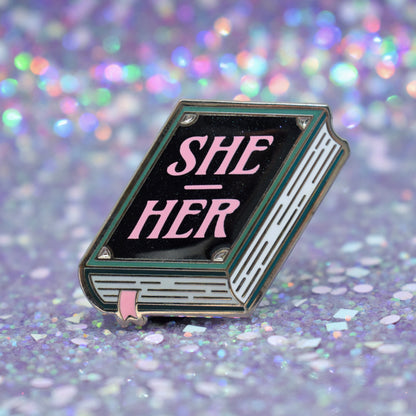 Book shaped enamel pin in silver metal, featuring teal, white and black shimmer accents with 'SHE - HER' in a light pink. Background is a glittery lilac.