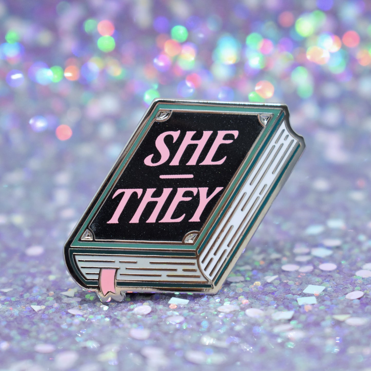 Book shaped enamel pin in silver metal, featuring teal, white and black shimmer accents with 'SHE - THEY' in a light pink. Background is a glittery lilac.