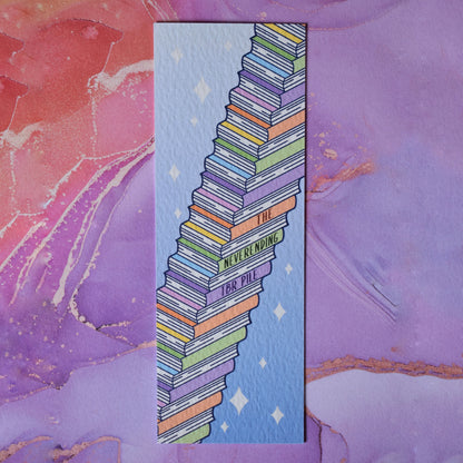 Textured bookmark with 'The neverending TBR pile' on an angled stack of books running from bottom left to top right. Sparkles on a gradient blue background fill the rest of the bookmark space. Background under bookmark is a peachy,lilac, gold watercolour style.