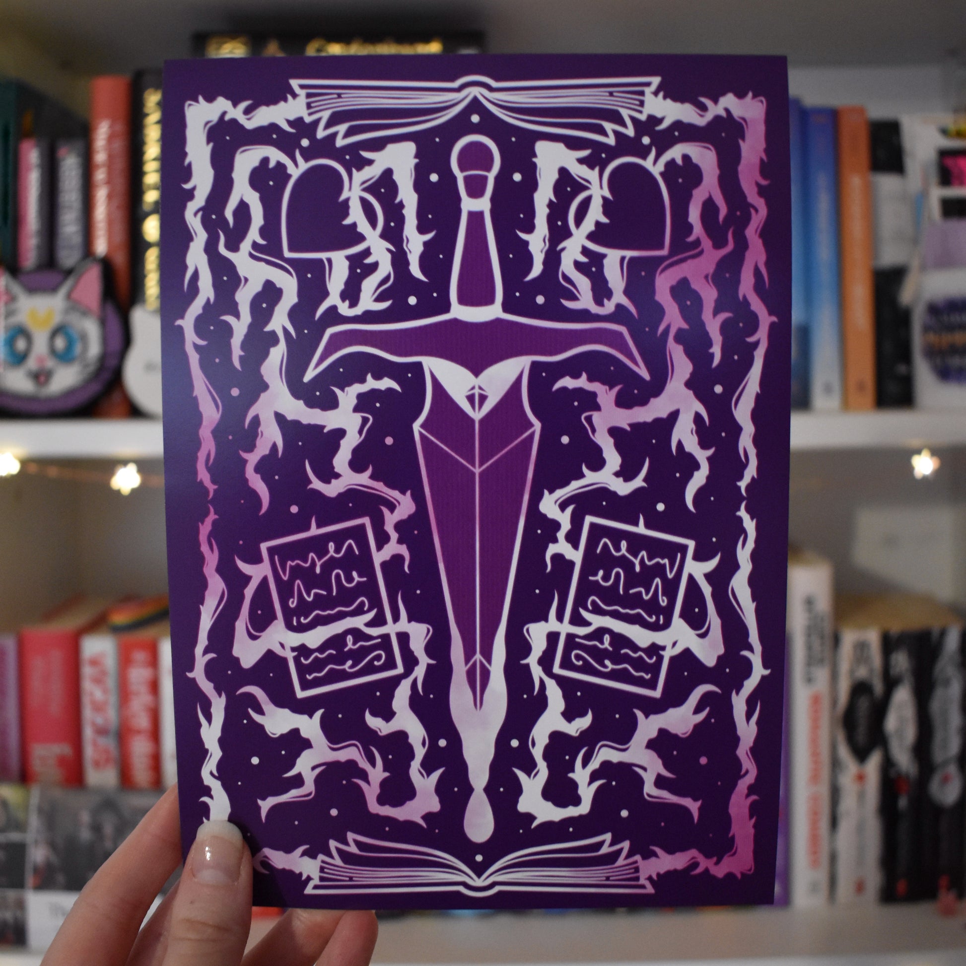 Purple illustrated design featuring white and pink, with a dagger in the centre, whispy/thunder/thorn branch elements mixed with open books, pages and hearts. Held by a white hand in centre of image against a blurry bookcase with fairy lights. 
