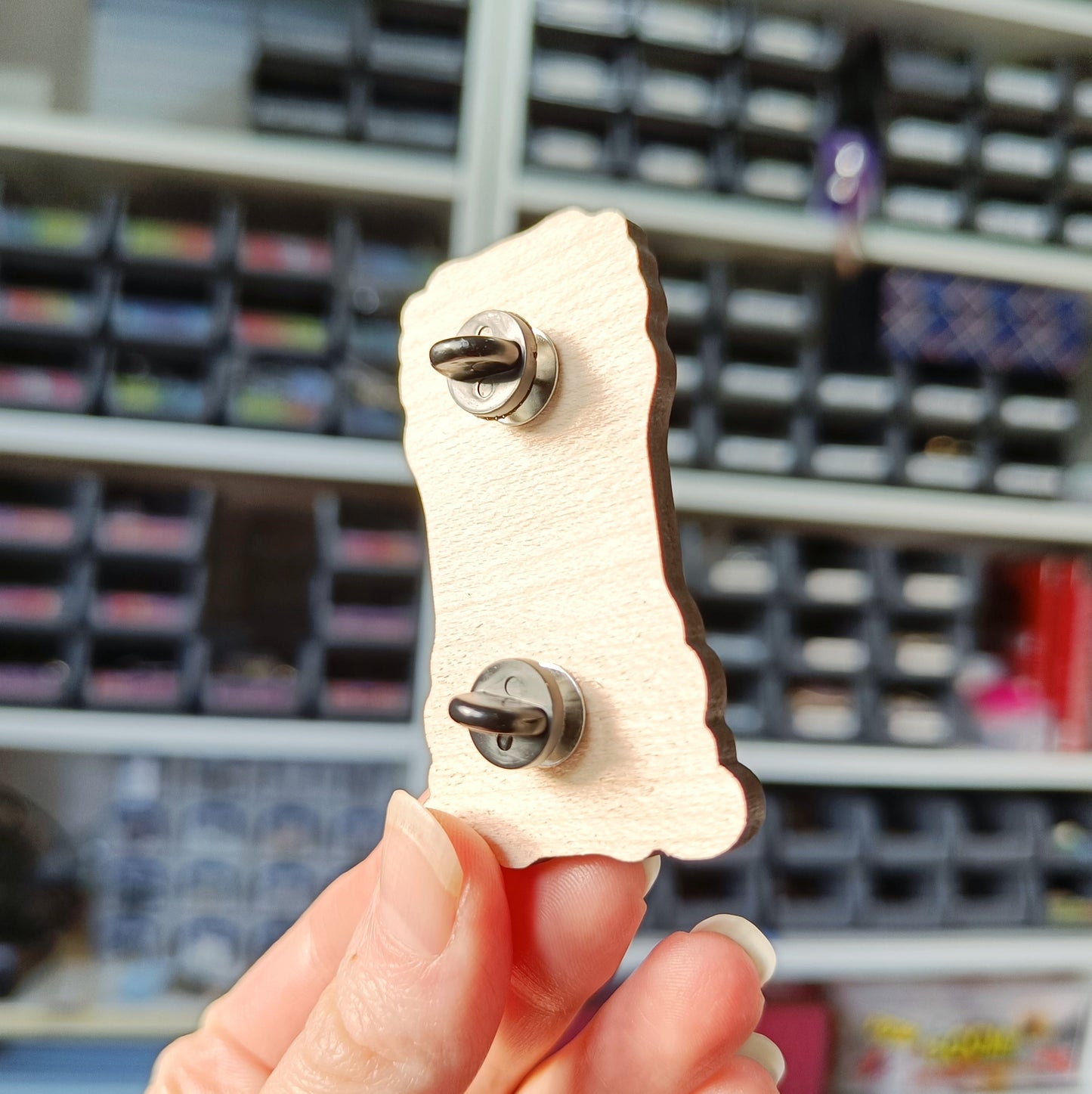 White hand holding wooden charm, showing the back of wooden charm to show placement of pin back option.