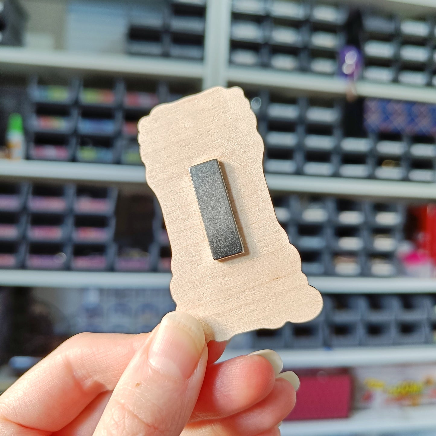 White hand holding wooden charm, showing the back of wooden charm to show placement of magnet option. Magnet is long silver bar lengthways down middle of charm.