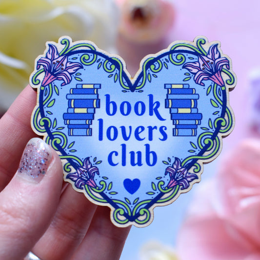 Blue heart-shaped bookish pin/magnet version, charm and keyring version, design features text 'book lovers club' with a floral and swirly outline with book spine stacks. Pin/magnet held by a white hand above a watercolour style background in blue, pink and purples with flowers around the edge of the photo.