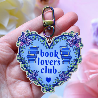 Blue heart-shaped bookish keyring version, charm and keyring version, design features text 'book lovers club' with a floral and swirly outline with book spine stacks. Keyring held by a white hand above a watercolour style background in blue, pink and purples with flowers around the edge of the photo.