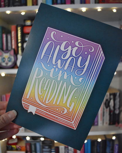 Teal illustration design with a rainbow book in the centre, facing forwards. 'Go away I'm reading' handwritten font in white ontop of the rainbow book. Held by a white hand at an angle in centre of image against a blurry bookcase with fairy lights.