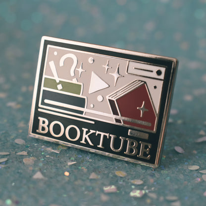 Silver metal enamel pin tilted to make the metal catch more light. Enamel pin is rectangular and black, with a smaller cream rectangle inside which has multicoloured books, sparkles, a !? and play button. Under the smaller rectangle says booktube with a couple decorative lines. Background of photograph is sparkly teal.