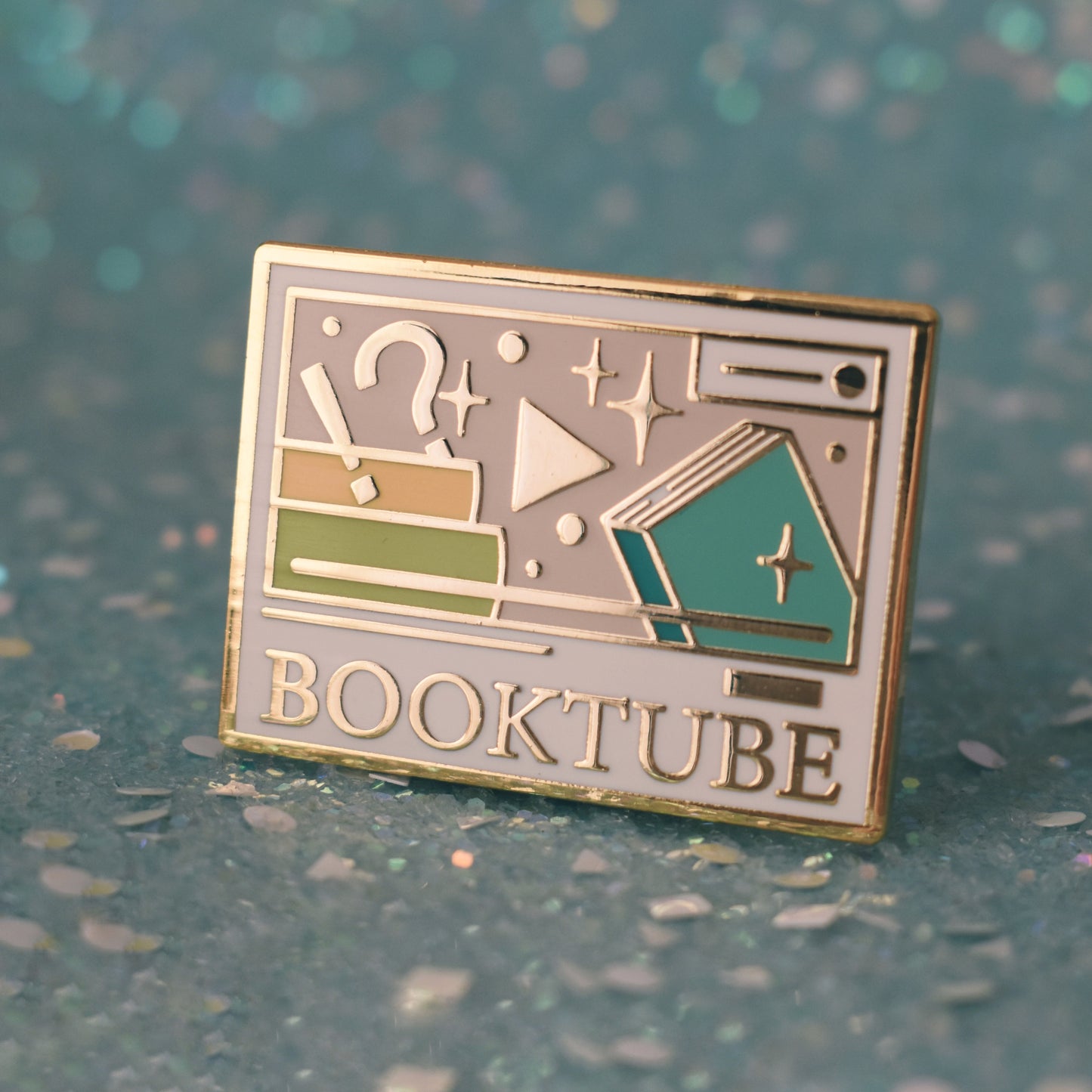 Gold metal enamel pin tilted to make the metal catch more light. Enamel pin is rectangular and white, with a smaller cream rectangle inside which has multicoloured books, sparkles, a !? and play button. Under the smaller rectangle says booktube with a couple decorative lines. Background of photograph is sparkly teal.